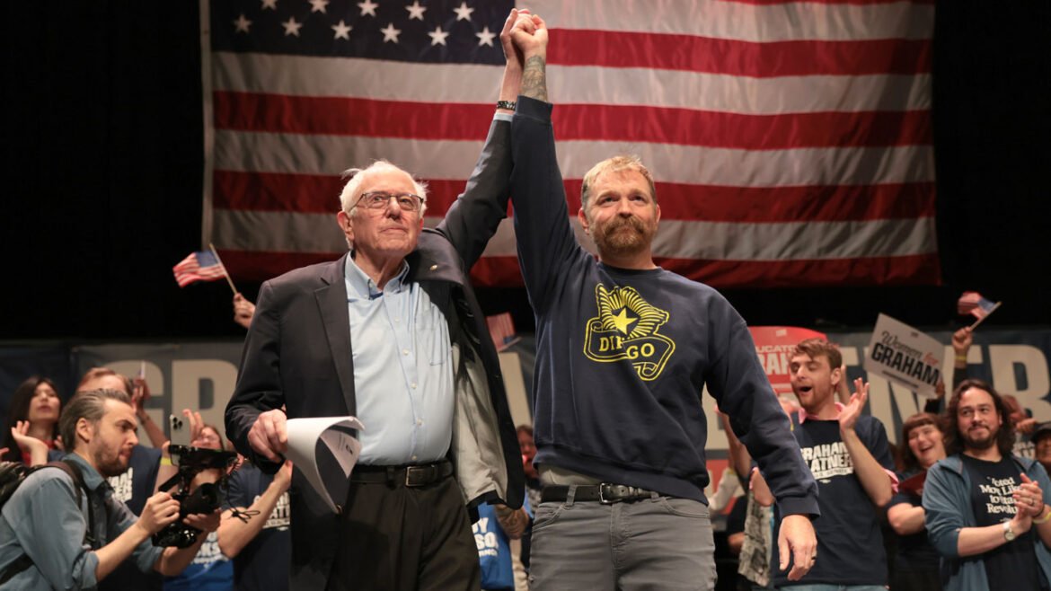 An older man in a suit holds up a mans arm on stage in front of a large American flag, with a cheering crowd in the background holding signs and waving flags.