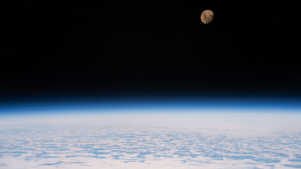 A view of the Moon from the International Space Station as it orbited 264 miles above a partly cloudy Indian Ocean southeast of Madagascar. The Moon has a golden glow to it, appearing more bronze than its usual gray. It stands out against the darkness of space, while below, Earth's atmosphere glows blue. Below that, we can see white clouds covering up most of the water below.