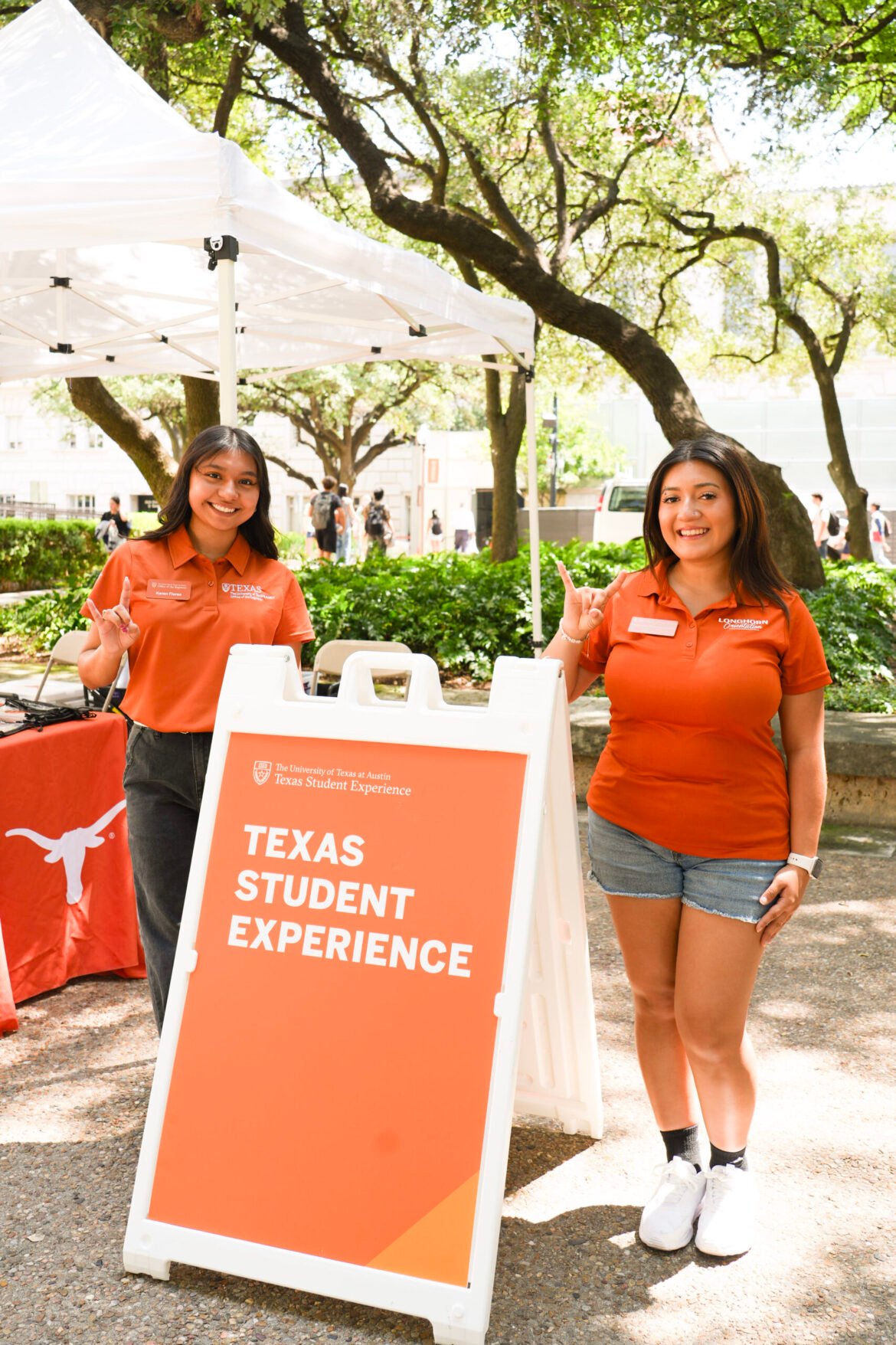 Two students in burnt orange pose outside under trees next to an orange sign