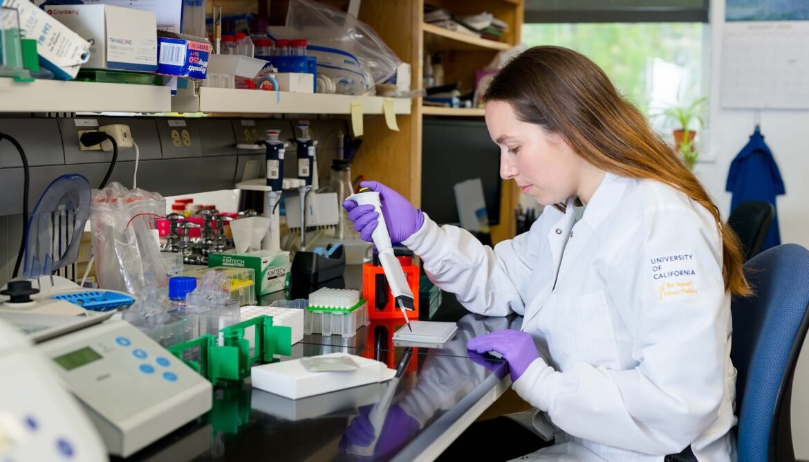 A person wearing a white lab coat and purple gloves operates a pipetting device on a lab bench.
