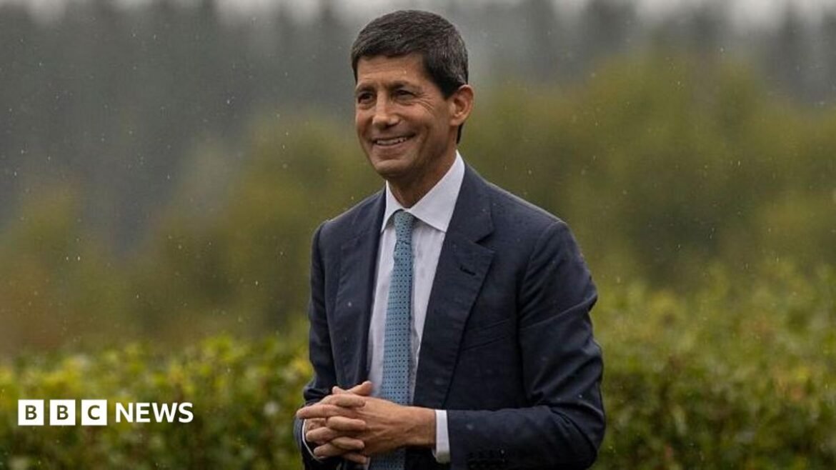 Federal Reserve Chairman Kevin Warsh at the Jackson Hole Economic Symposium in Wyoming. He is wearing a dark suit with a white shirt and blue-spotted tie. He has his hands clasped together and is smiling.  