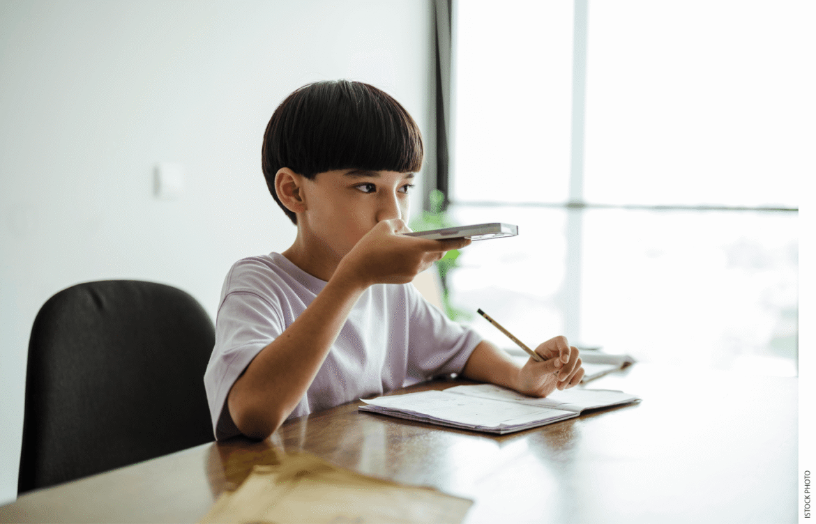 A boy, aged 8 or 9, sits a table at home with an open notebook in front of him, a pencil in his left hand, and a mobile phone in his right hand. He is holding the phone horizontally flat up to his mouth and speaking into one end of it.