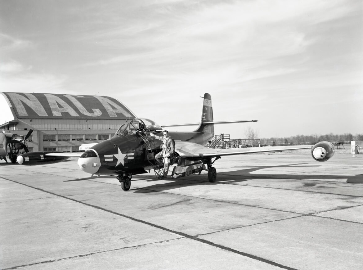 A pilot steps on a metal ladder to get onto a small, one-seater airplane. The NACA hangar is in background