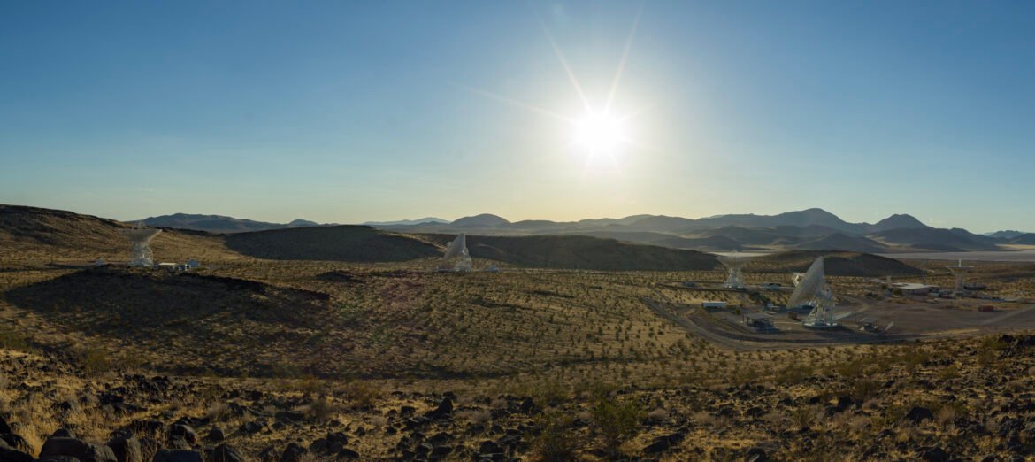 A wide desert landscape featuring several large white satellite dishes pointing toward a bright sun shining in a clear blue sky above distant mountain ranges.