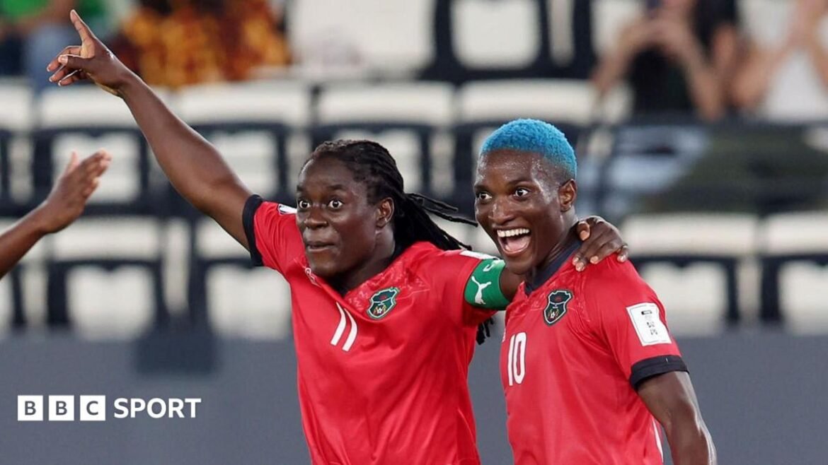 Tabitha Chawinga raises her right arm in celebration and puts her left arm around the shoulder of Temwa Chawinga, who has dyed blue hair and a wide-mouthed smile on her face. Both players wear red Malawi shirts with black trim - Tabitha with a green captain's armband and a white number 11 on the front and Temwa with a number 10 on the chest. The pair are visible from stomatch up and a sparsely-populated stand can be seen out of focus in the background 
