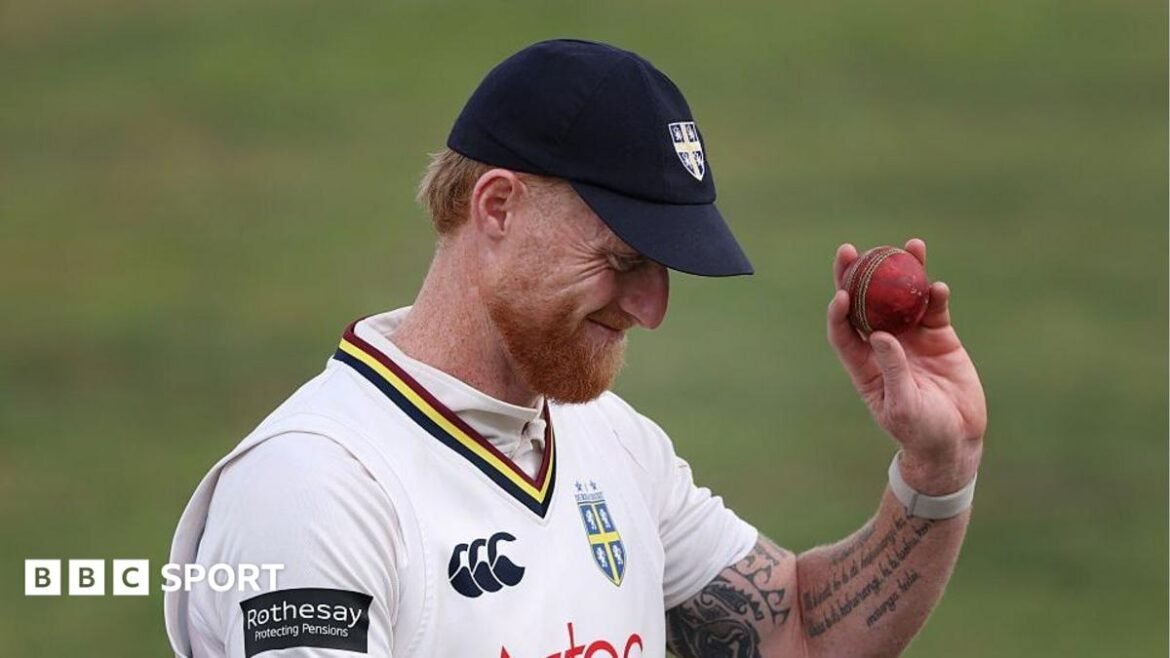 Ben Stokes raises the red ball in his left hand as he bows his head, while wearing his navy Durham cap