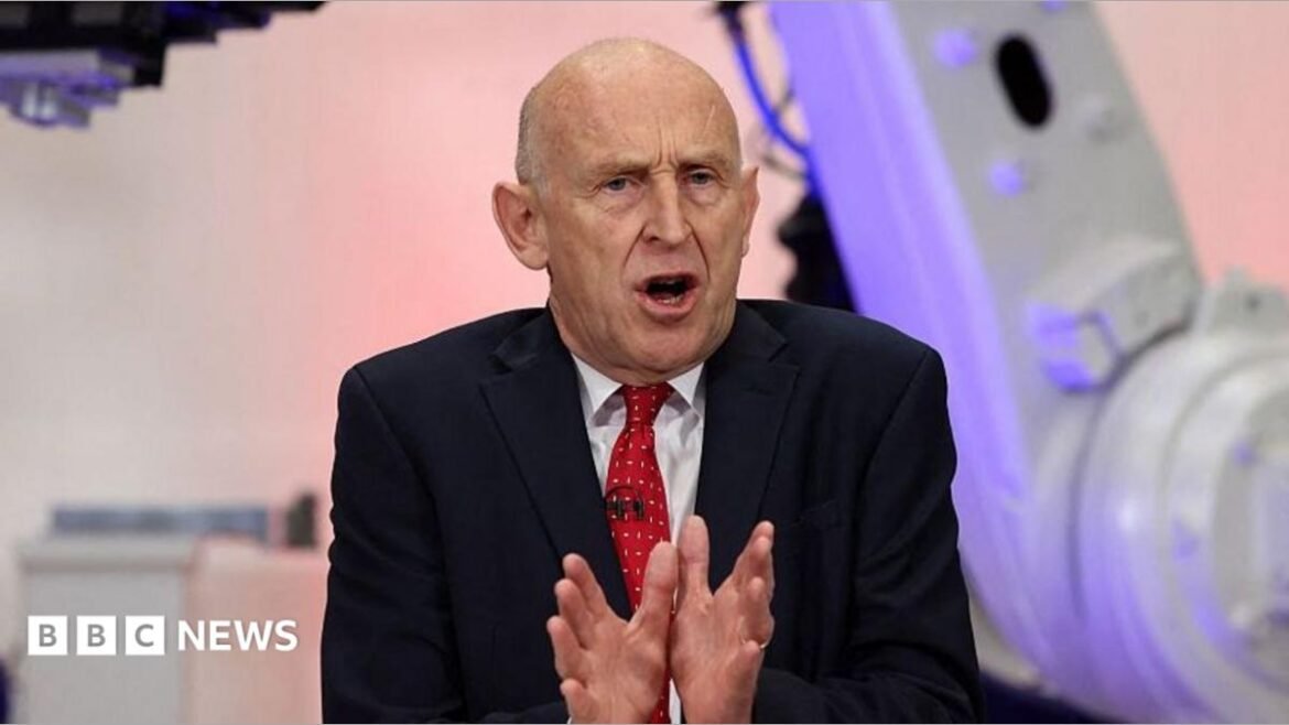 Chancellor John Healey speaking in Coventry in front of purple and grey coloured equipment. He is wearing a dark suit, white short and red tie 