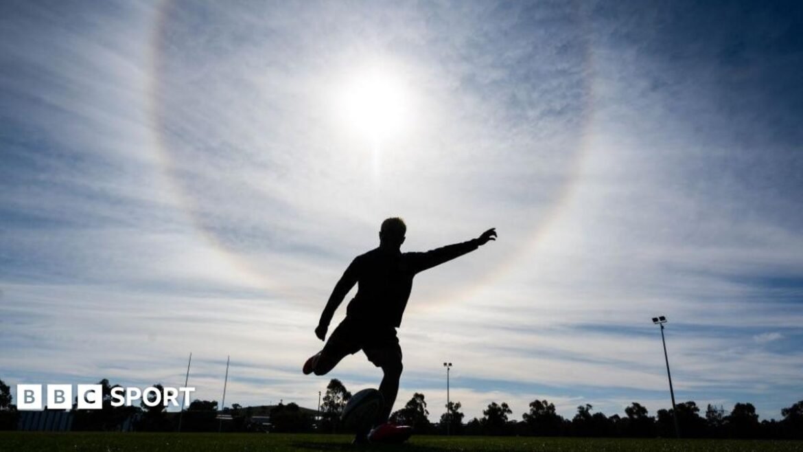 A silhouetted rugby player strikes the ball off a tee during a training session, with the sun glaring down from behind him