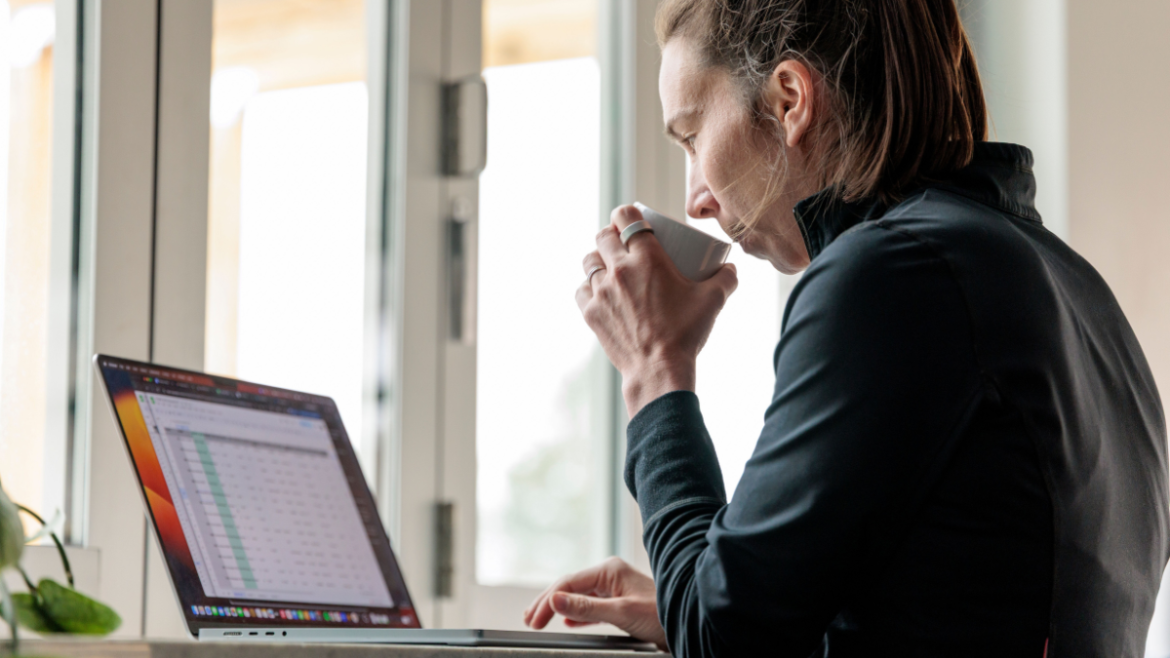 Coach Drinking Coffee Out Of White Mug And Forming Athlete Training Plans On Her Laptop Using Her Decision Making Framework.
