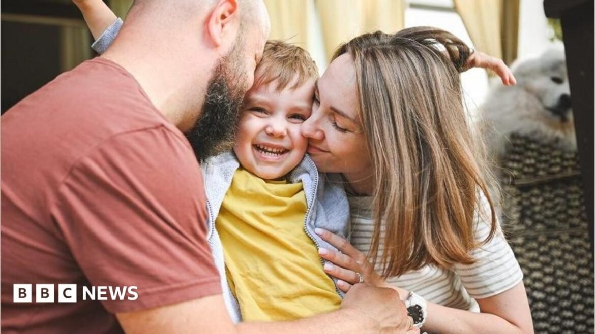 A young blonde child wearing a yellow t-shirt is hugged by a man wearing a red t-shirt and a woman wearing a beige striped t-shirt.