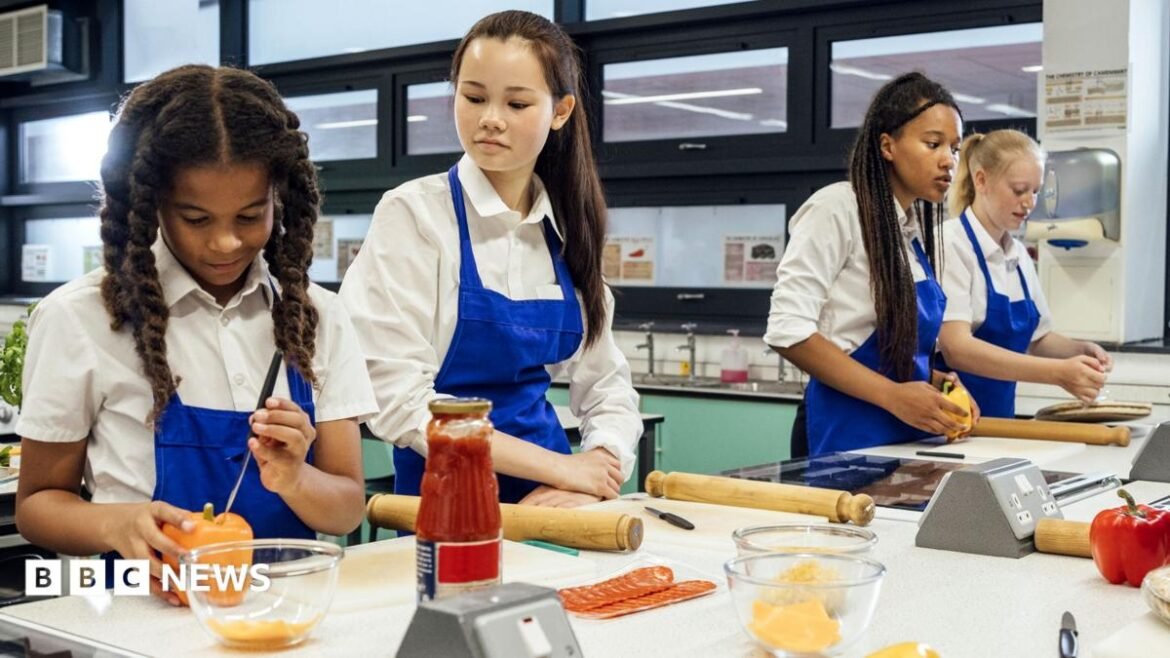Two teenagers at a classroom bench, wearing aprons., They are in a catering class and are adding cream cheese to bowls, and are being supervised by their female teacher, also in an apron, who is standing to the left of them. 