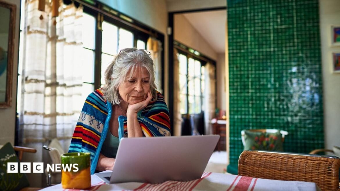 A pensioner looks at her finances on her laptop at her dining room table. She is wearing a colourful throwover.
