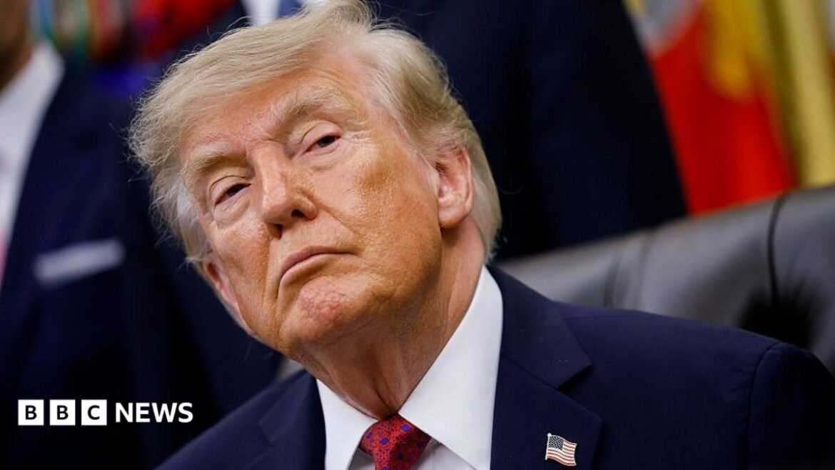 US President Donald Trump sitting down in the Oval Office at the White House. He is wearing a dark blue suit, white shirt and a red tie with a blue pattern on it.