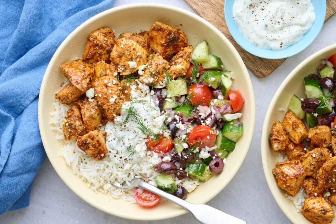 Two Greek chicken bowls served over rice with chicken, veggie salad, and tzatziki segmented on top, garnished with feta and fresh dill with a dish of more tzatziki nearby.