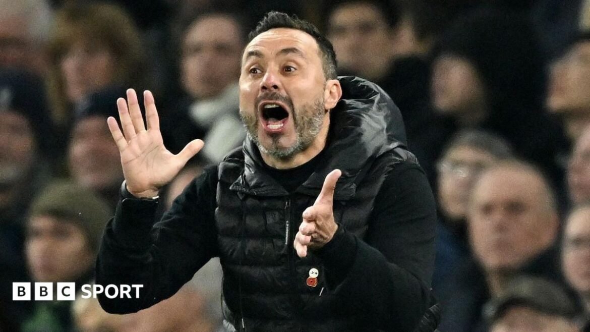 Tottenham Hotspur head coach Roberto de Zerbi looks animated with his mouth wide open and both hands out in front of him as if about to applaud, as he wears a black padded jacket, which has a poppy badge attached