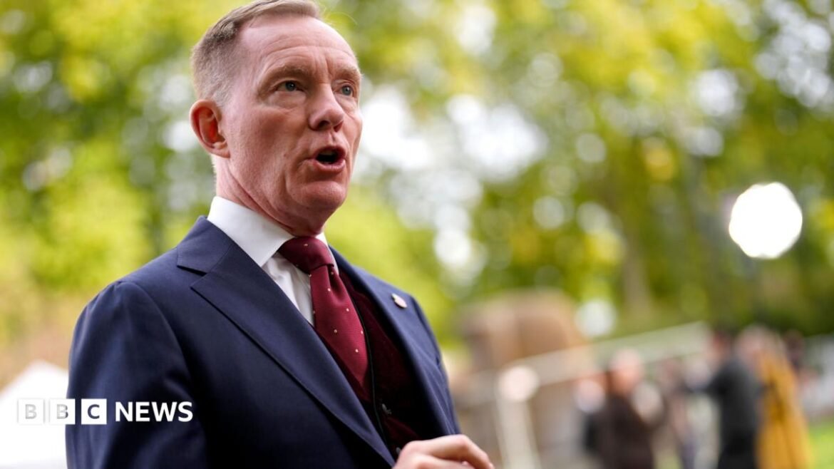 Chris Bryant is standing to the side, mouth open mid-talking. There are blurred trees and figures behind him. He is wearing a blue three-piece suit and red tie with white dots.