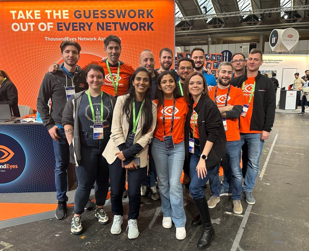 A group of colleagues poses together at a trade show booth featuring ThousandEyes and Cisco branding, with a bright orange backdrop displaying the message “Take the guesswork out of every network.” Several team members wear orange ThousandEyes T-shirts and conference badges.