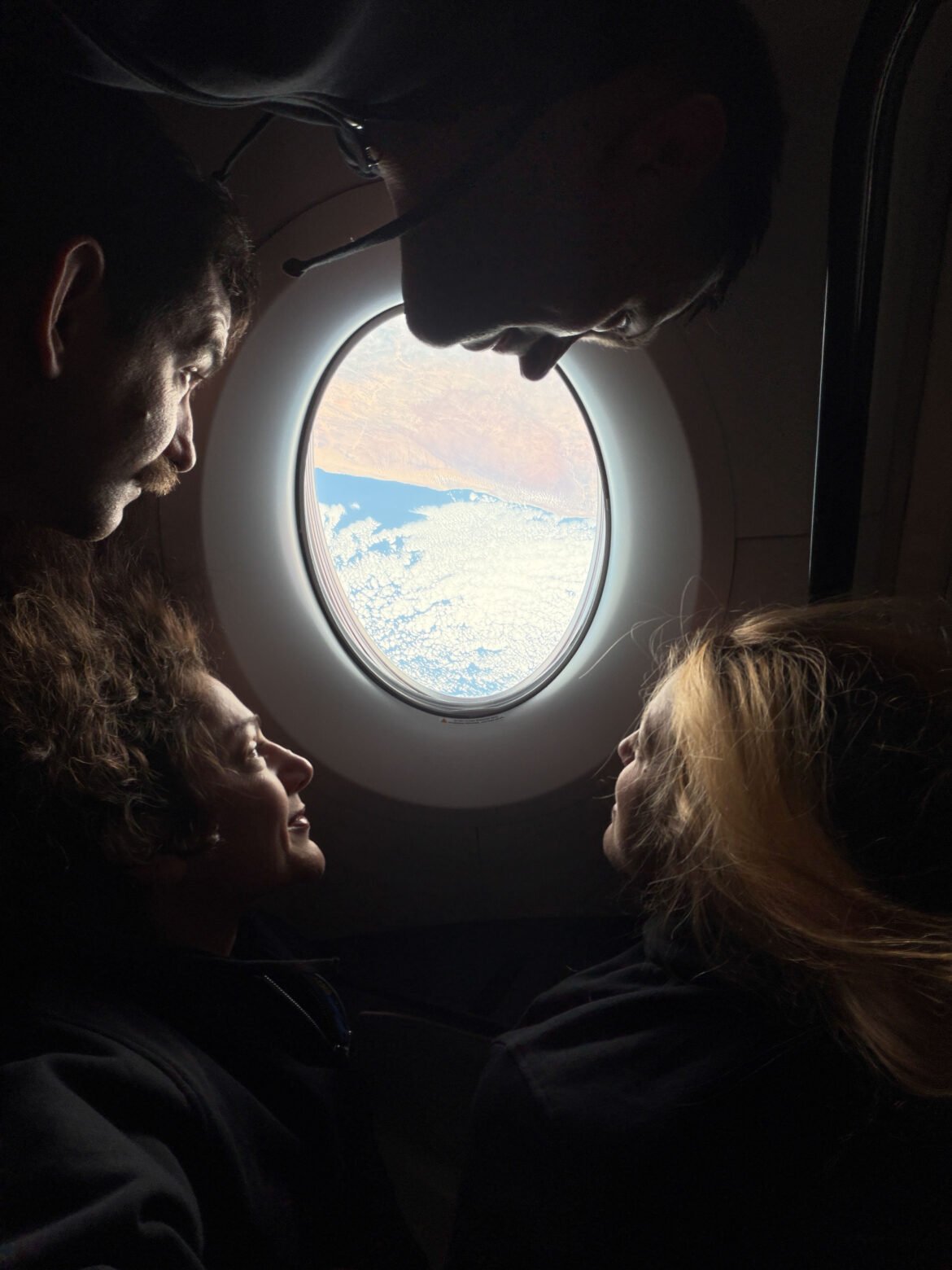 NASA’s SpaceX Crew-12 crew members surround an oval-shaped window aboard a SpaceX Dragon, looking out at clouds above blue water and a tan landmass. From the left, rotating clockwise around the image are NASA astronaut Jack Hathaway, Roscosmos cosmonaut Andrey Fedyaev, ESA (European Space Agency) astronaut Sophie Adenot, and NASA astronaut Jessica Meir. Their faces are illuminated by sunlight streaming through the window.