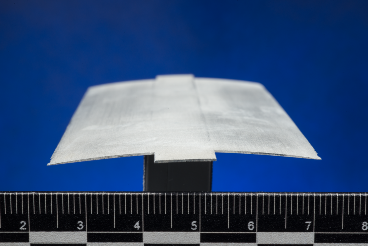 A close-up view of a cross section of a rotor blade, also called an airfoil. The thin, gray material is slightly arched and sits atop a black stand against a ruler.