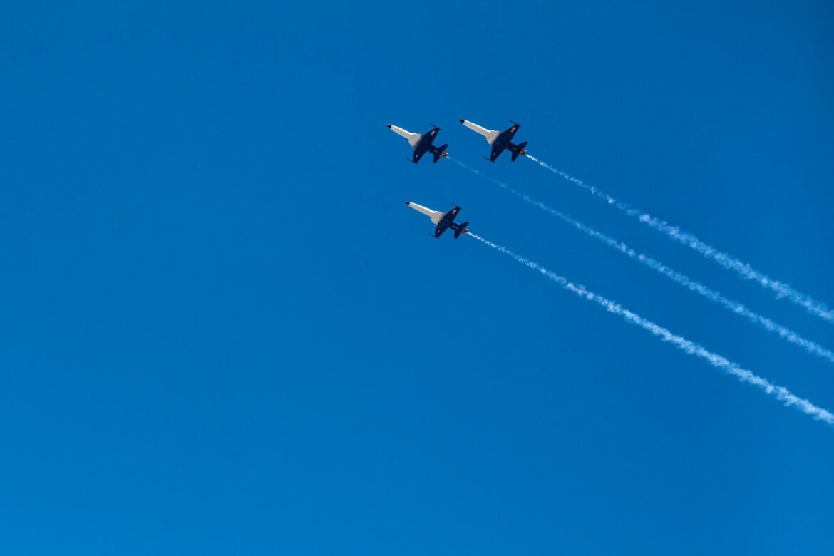 Looking up, three white and black fighter jets fly in formation across a bright, blue sky.