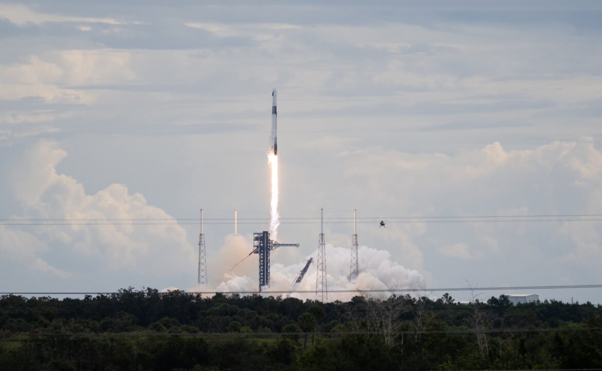 A SpaceX Falcon 9 rocket carrying the company's Dragon spacecraft launches. There is a bright column of fire under the rocket as it moves upward. Clouds of vapor spread out below the rocket, almost blending into the similarly shaped clouds in the sky.