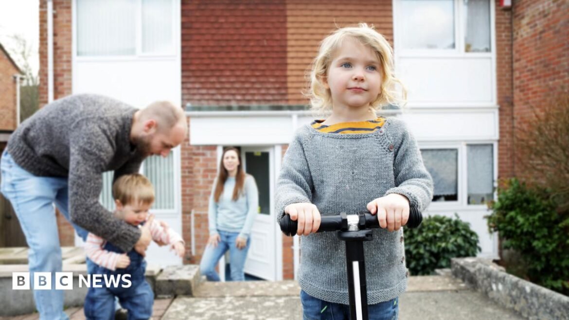 Family outside a house including a man, woman with a small child on her hip and another small child riding a scooter in the foreground