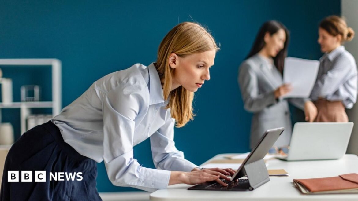 A female office worker wearing a light blue top and dark blue skirt leans over a laptop computer. Two other workers are in the background, slightly out of focus