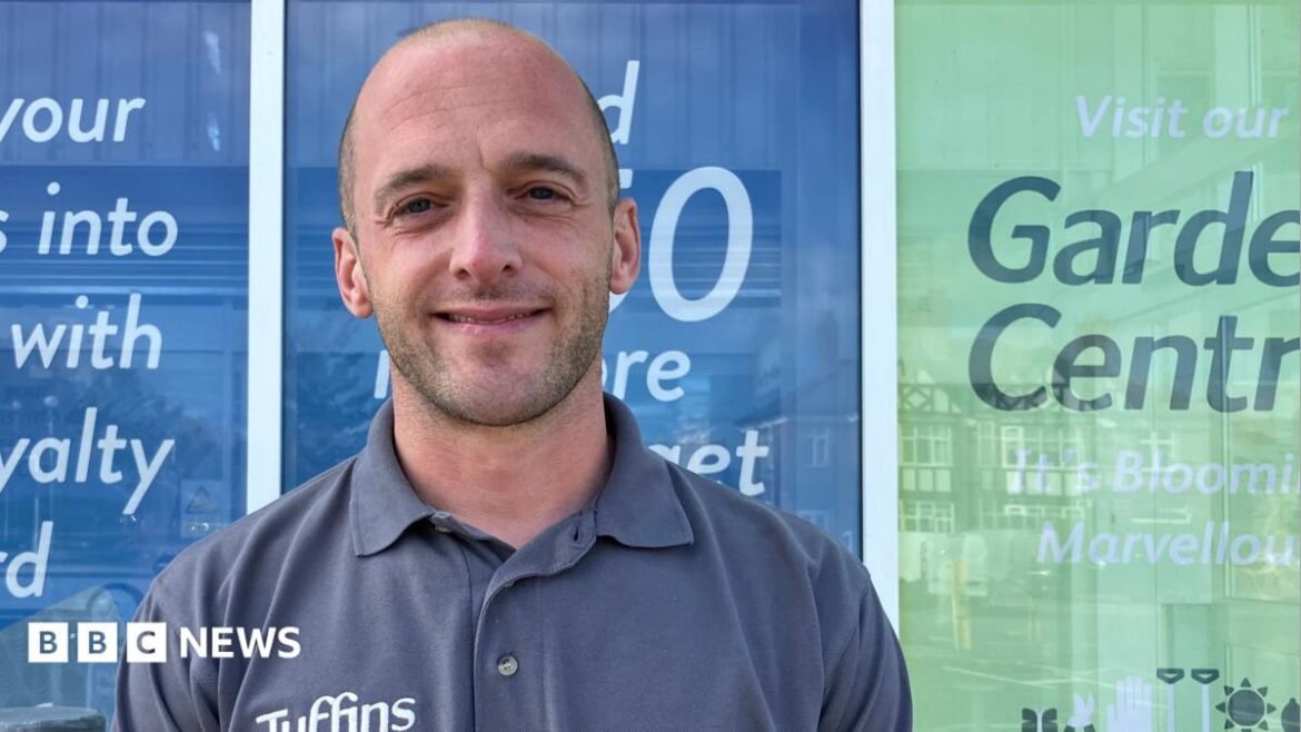 A person wearing a grey Tuffins Supermarket polo shirt stands outside a shopfront with promotional signage in the background.